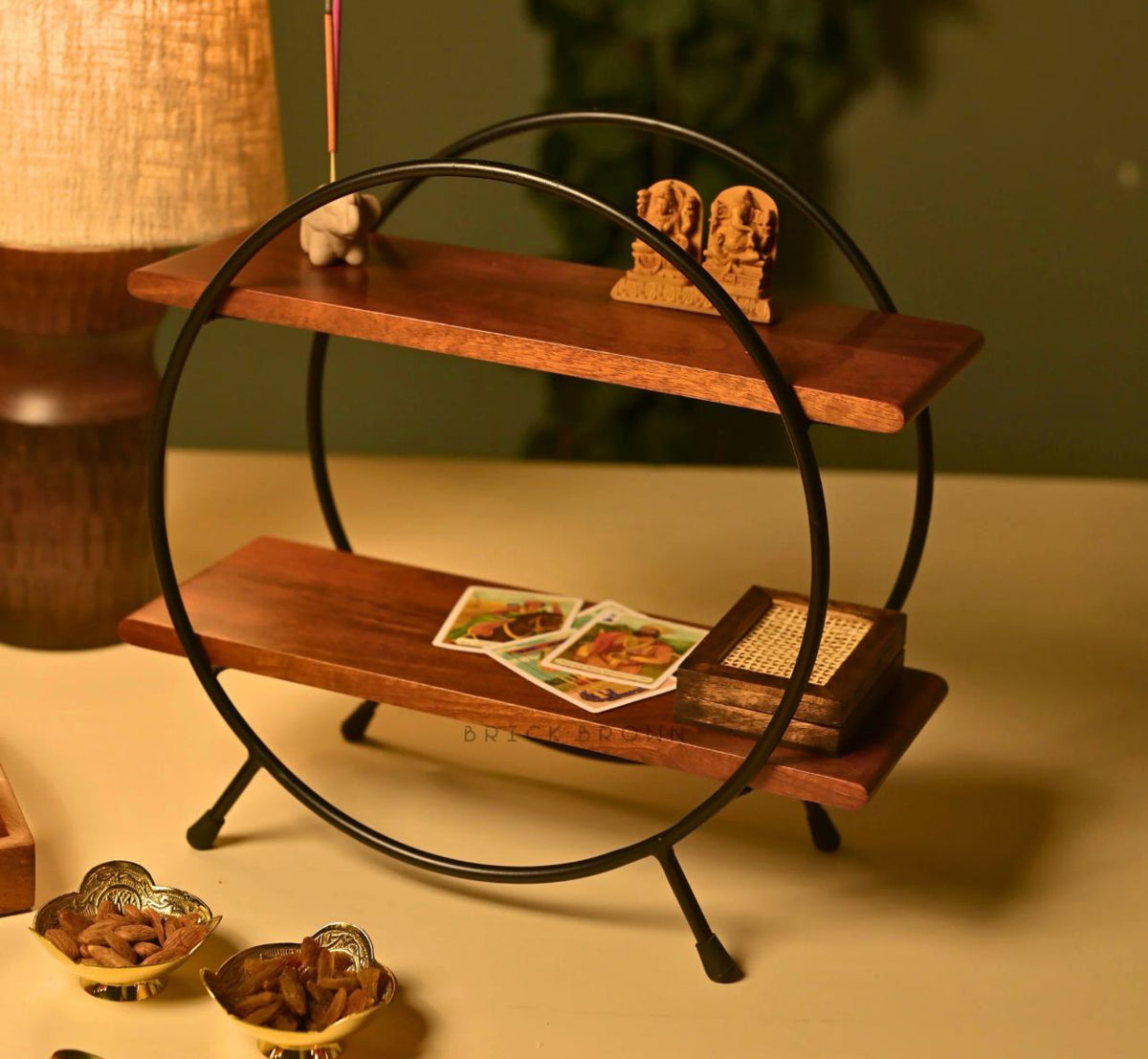 Orbit Duo Shelf by Metallics Home features a circular design with two wooden shelves and black metal legs, ideal for decor.