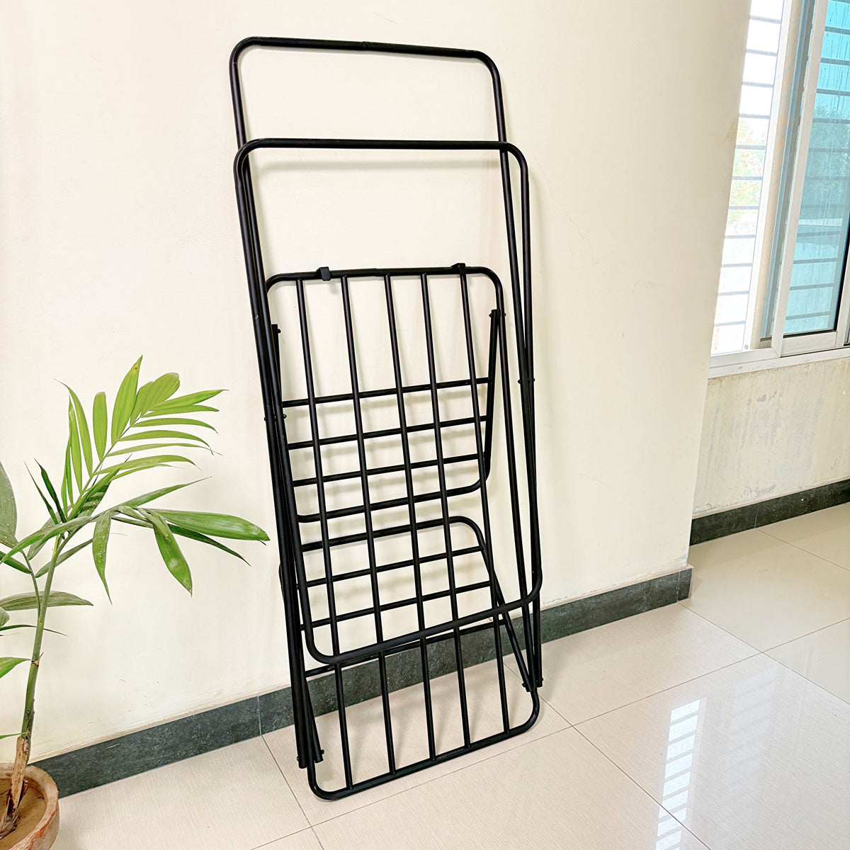Premium Metal Folding Cloth Dryer Stand by Metallics Home, black, compact design, leaning against a wall with a plant nearby.